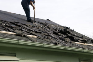 Roofer is removing shingles from atop a roof during the day.